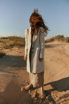 a woman poses standing on the sand in light summer clothes against the blue sky and the wind blows her hair on her face. High quality photo