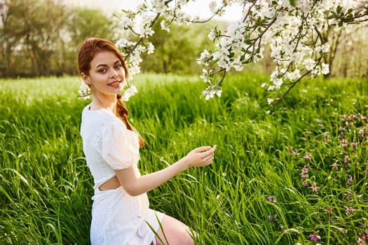 a lovely woman in a light short dress sits near a flowering tree and smiles while looking at the camera. High quality photo