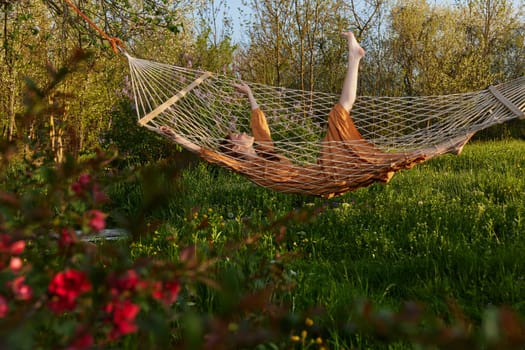 a happy woman in a long orange dress is relaxing in nature lying in a mesh hammock enjoying summer and vacation in the country surrounded by green foliage, happily lifting her legs. High quality photo