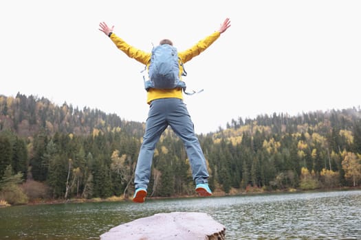 Happy male traveler with backpack jumping near forest lake while reaching goal back view closeup. Freedom and fulfillment of desires concept