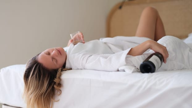 Drunk woman in bath robe lying in bed with bottle of wine in hotel. Loneliness and alcohol addiction in women concept