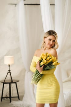 a happy woman in a yellow dress embraces a bouquet of yellow spring tulips in the interior.