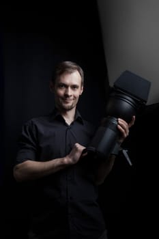 Portrait of a gaffer man with a professional lighting head device in his hands on a black background.