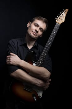 Young man low key portrait hugging electric guitar look at camera on black background.