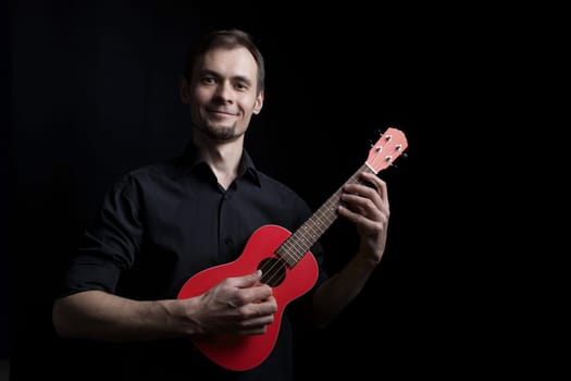 Handsome smiling low key man portrait playing on fun red ukulele guitar over black background.