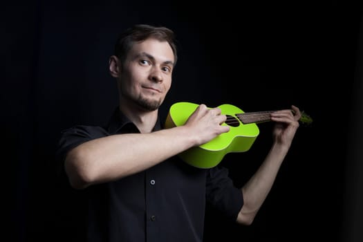 Handsome young low key man portrait with fun green ukulele guitar over black background.