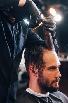 Young bearded man sitting and getting haircut in barber shop by guy in black protective mask.