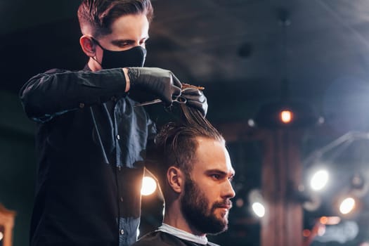 Young bearded man sitting and getting haircut in barber shop by guy in black protective mask.