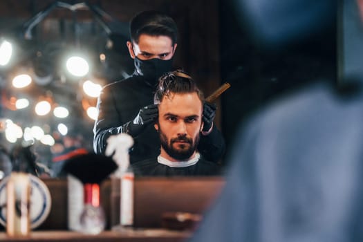 Front view of young bearded man that sitting and getting haircut in barber shop by guy in black protective mask.