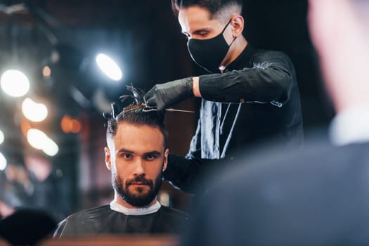 Front view of young bearded man that sitting and getting haircut in barber shop by guy in black protective mask.