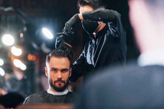 Front view of young bearded man that sitting and getting haircut in barber shop by guy in black protective mask.