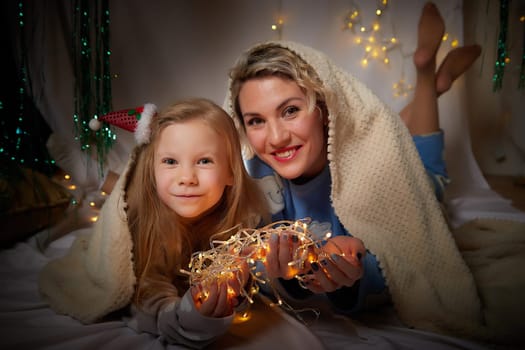 Cute mother and daughter in pajamas lie on soft blanket and having fun in room with Christmas garlands and white background. Tradition of decorating house for holiday. Happy childhood and motherhood