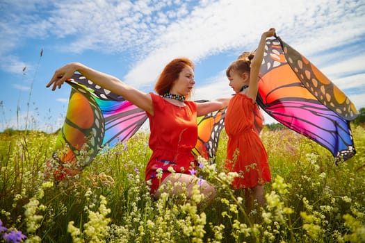 Happy female family with red haired mother and daughter with bright butterfly wings having fun on green and yellow meadow full of grass and flowers in sunny summer day. Concept family love