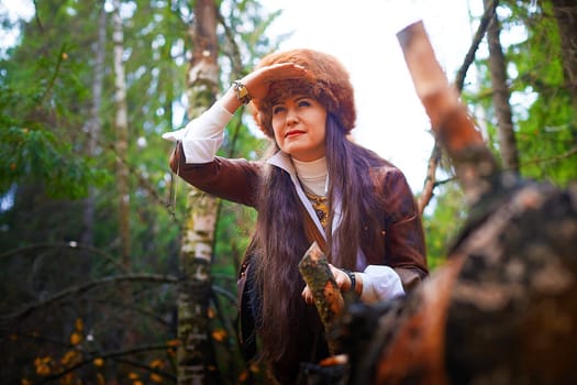 Girl in a leather jacket, a big red fox fur hat in the forest in autumn. A female model poses as a fabulous royal huntress on nature hunt at a photo shoot