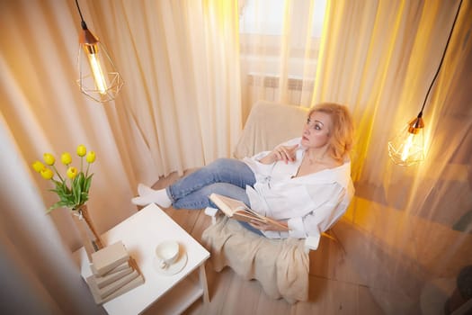 Adult mature woman of 40 or 60 years old with book in casual dress white shirt and blue jeans in calm cozy evening atmosphere room. Interior with transparent curtains and soft warm lamp