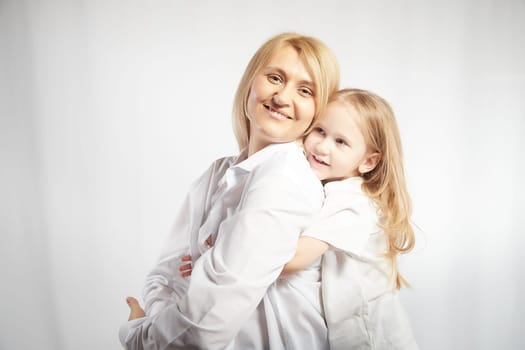 Close portrait of a blonde mother and daughter with hugs on white background. Mom and little girl models pose in the studio. The concept of love, friendship, caring in the family