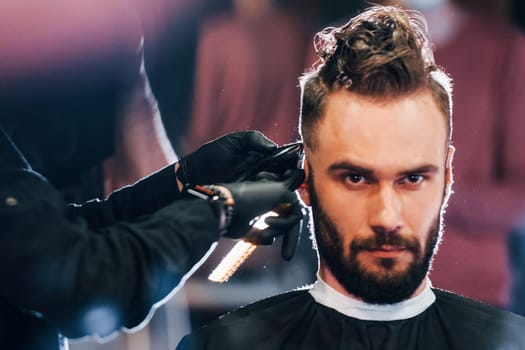 Young bearded man sitting and getting haircut in barber shop.