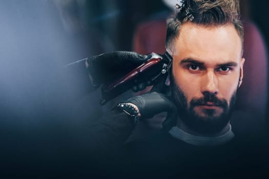 Young bearded man sitting and getting haircut in barber shop.