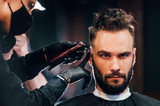 Front view of young bearded man that sitting and getting haircut in barber shop by guy in black protective mask.