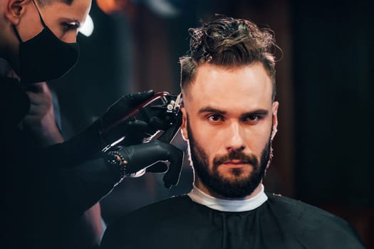 Front view of young bearded man that sitting and getting haircut in barber shop by guy in black protective mask.