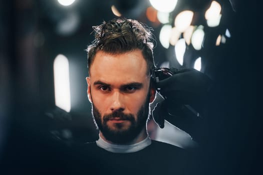 Young bearded man sitting and getting haircut in barber shop.