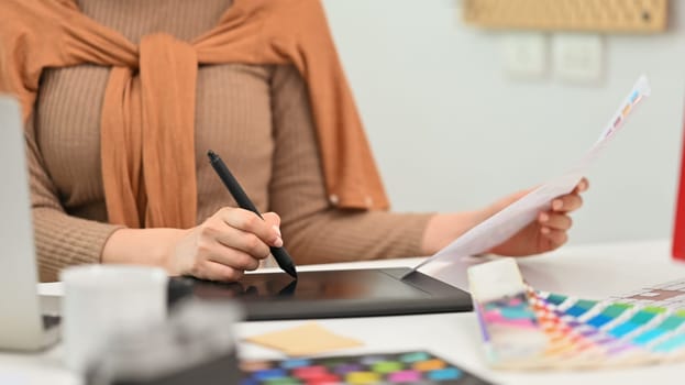 Cropped shot, selective focus on hand. Creative woman using graphic tablet and working with color swatch samples at workplace.