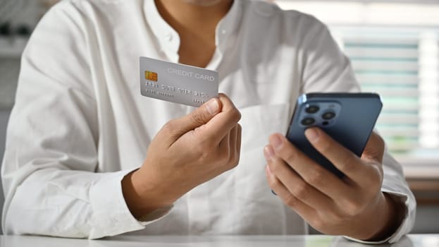 Cropped image of man holding using mobile phone and credit cad for online banking transaction, ordering in internet.