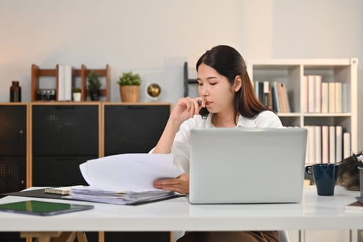 Concentrated female siting in front of laptop computer at working desk and checking marketing reports.