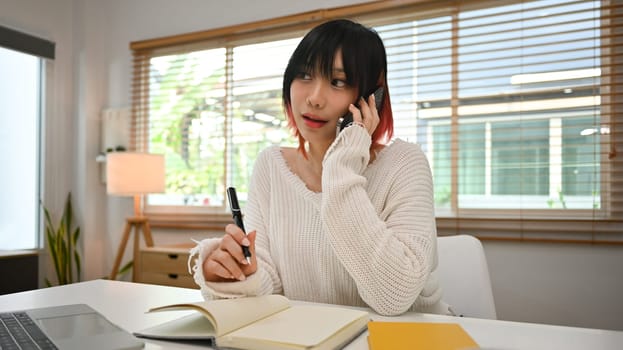 Charming young Asian woman freelancer having phone conversation and making note on notebook.
