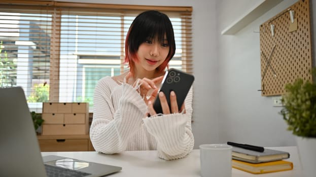 Beautiful young asian woman in casual clothes sitting in home office checking social media, chatting online on smartphone.