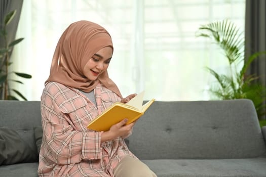 Happy young muslim woman wear hijab and casual clothes reading book on couch, spending time at home.
