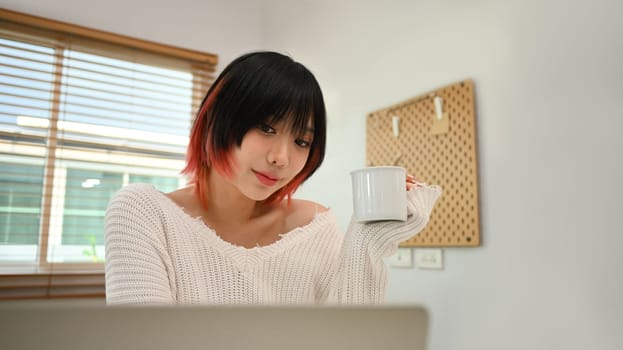Pretty young asian woman in casual clothes drinking hot coffee and browsing internet on laptop at cozy home.