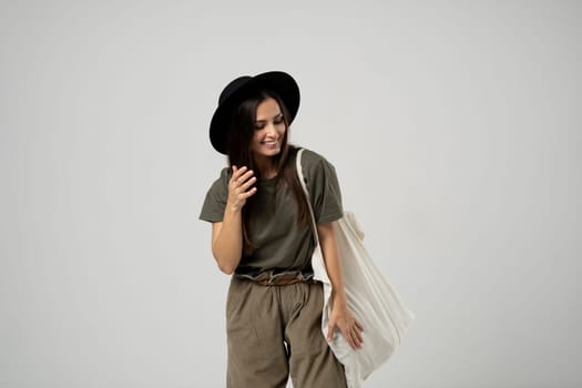 Brunette woman in green t-shirt and black hat holding cotton shopper bag with vegetables, products in white room. Eco friendly shopping bags. Zero waste, plastic free concept