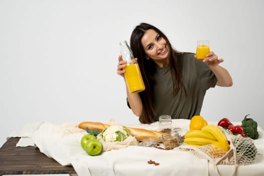 Happy woman in green t-shirt unpacking shopping mesh eco bag with healthy vegetables, fruits, bread, snacks on the kitchen at home. Healthy eating vegetarian concept