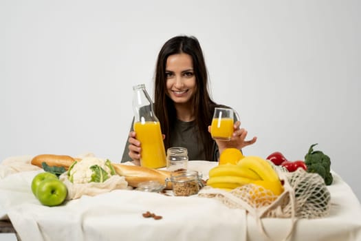 Woman in green t-shirt pouring a juice from a glass bottle in a glass over a table with mesh eco bag, healthy vegan vegetables, fruits, bread, snacks. Zero waste concept