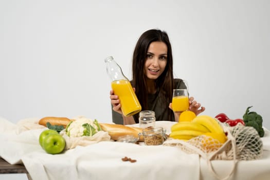 Woman in green t-shirt drink a juice over a table with mesh eco bag, healthy vegan vegetables, fruits, bread, snacks. Zero waste concept