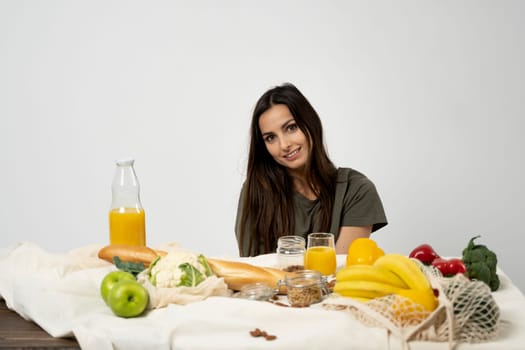 Happy woman in green t-shirt unpacking shopping mesh eco bag with healthy vegetables, fruits, bread, snacks on the kitchen at home. Healthy eating vegetarian concept