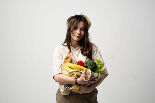 Smiling young woman in light summer clothes with a mesh eco bag full of vegetables, greens watching in a camera on a green studio background. Sustainable lifestyle. Eco friendly concept. Zero waste