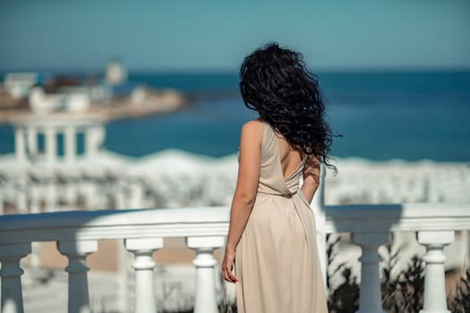 Sea woman rest. A woman with long curly hair in a beige dress stands with her back and looks at the sea and the coast from a balcony with balusters. Tourist trip to the sea