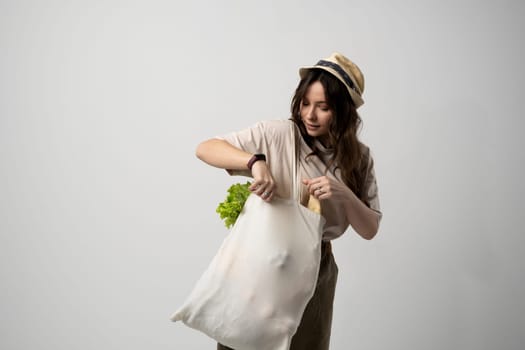 Smiling woman in a t-shirt and a hat holding reusable cotton shopping bag with vegetables, bread and greens. Concept of no plastic, zero waste, plastic free, sustainable lifestyle