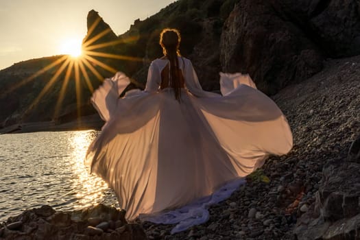 Mysterious woman silhouette long hair walks on the beach ocean water, sea nymph wind listens to the wave. Throws up a long white dress, a divine sunset. Artistic photo from the back without a face.