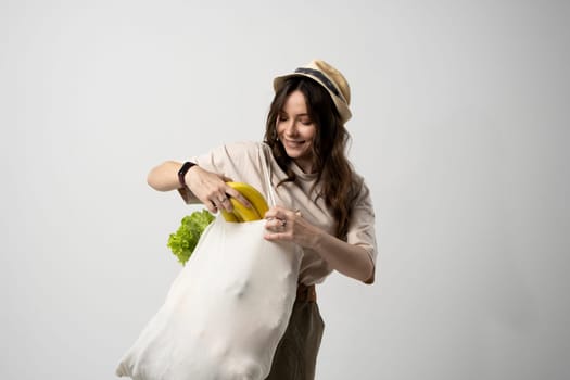 Smiling young woman in light summer clothes with a mesh eco bag full of vegetables, greens watching in a camera on a green studio background. Sustainable lifestyle. Eco friendly concept. Zero waste