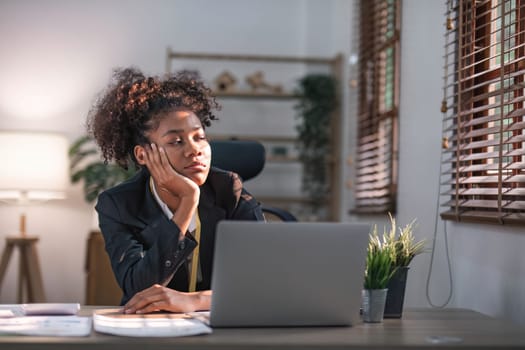 Young African American woman with afro hairstyle looks annoyed and stressed, sitting at the desk, using a laptop, thinking and looking at the camera, feeling tired and bored with depression problems..