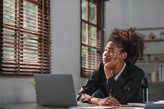 Young African American woman with afro hairstyle looks annoyed and stressed, sitting at the desk, using a laptop, thinking and looking at the camera, feeling tired and bored with depression problems..