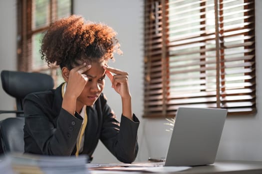 Young African American woman with afro hairstyle looks annoyed and stressed, sitting at the desk, using a laptop, thinking and looking at the camera, feeling tired and bored with depression problems..