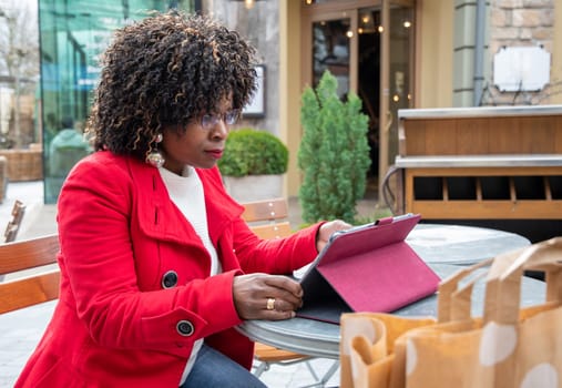 a young african american woman in a red coat sits at a table in a cafe and checking bill after shopping using a tablet, financial problems crisis, proper planning of expenses, High quality photo