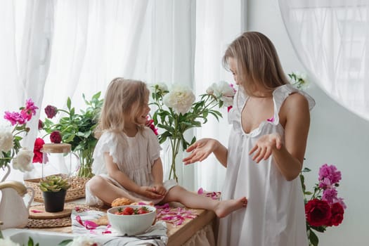 A little blonde girl with her mom on a kitchen countertop decorated with peonies. The concept of the relationship between mother and daughter. Spring atmosphere