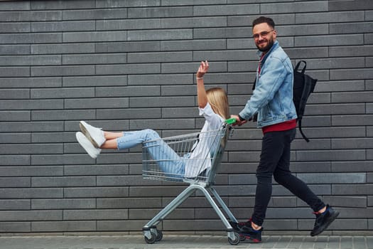 Having fun and riding shopping cart. Young stylish man with woman in casual clothes outdoors together. Conception of friendship or relationships.