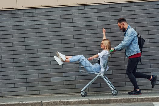 Having fun and riding shopping cart. Young stylish man with woman in casual clothes outdoors together. Conception of friendship or relationships.