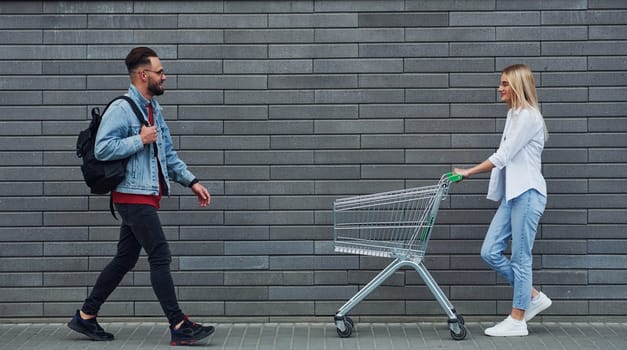 With shopping cart. Young stylish man with woman in casual clothes outdoors together. Conception of friendship or relationships.